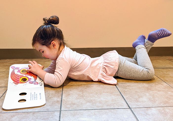 A young child is lying on her tummy on the floor, looking at a picture book in front of her