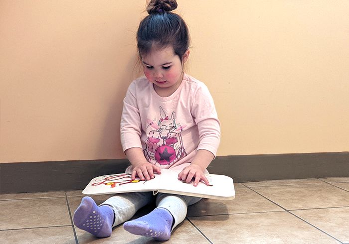 A young child is reading a book while sitting on the floor with her back supported by a wall and her legs sticking out straight