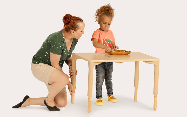 A young child is standing at a table while a teacher adjusts the table height