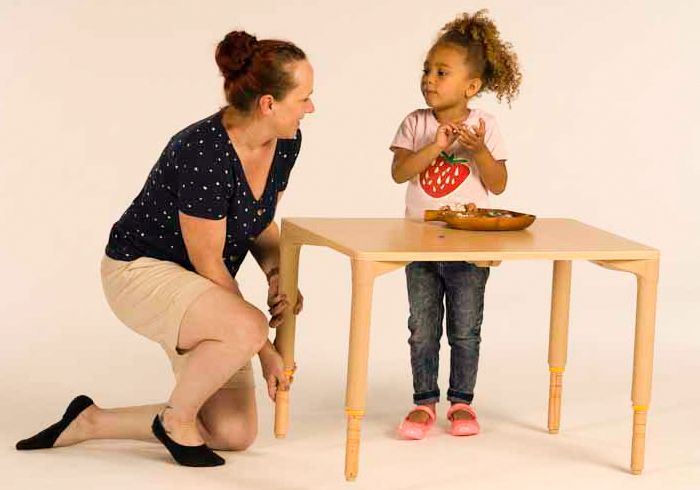 A young child is standing at a table while a teacher adjusts the table height