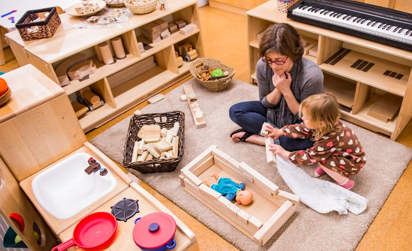 A child and a teacher sitting on the floor role-playing with wooden blocks and a doll.