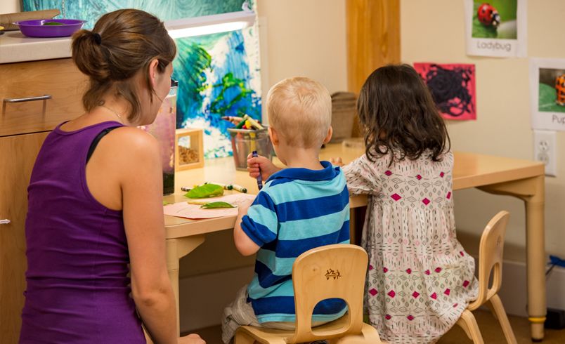 A teacher is looking at two children sitting at a table with crayons and art materials.