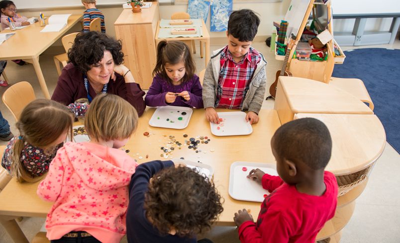 A group of children and a teacher are sitting or standing around a table playing with coloured glass beads.